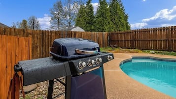 A barbecue grill with a lid, a pool, and a wooden fence in the background.
