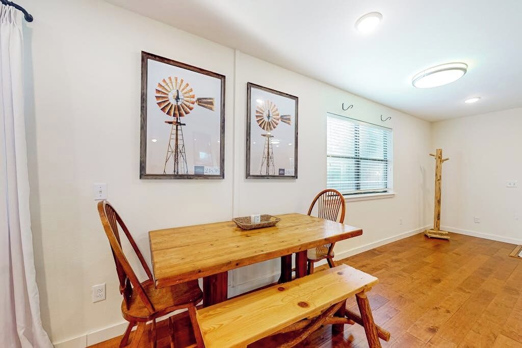 A dining area with a wooden table and benches, framed windmill artwork on the wall, and a window with blinds.
