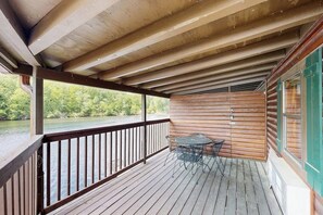 Eine Holzdar Terrasse mit Geländer, einem Tisch mit Stühlen und Blick auf einen Fluss und Grünflächen.