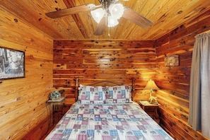 Une chambre dans une cabane en bois, avec un lit, un ventilateur de plafond, une table de chevet avec une lampe et un tableau encadré au mur.