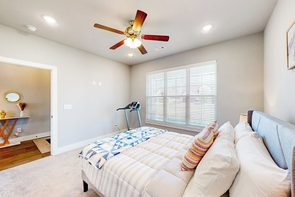 A bedroom with a bed, a ceiling fan, a treadmill, and a window with blinds.