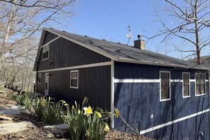 A single-story building with a dark facade and white trim, surrounded by bare trees and daffodils. - 4BR Waterfront - Screen-in Deck (Eureka Springs)