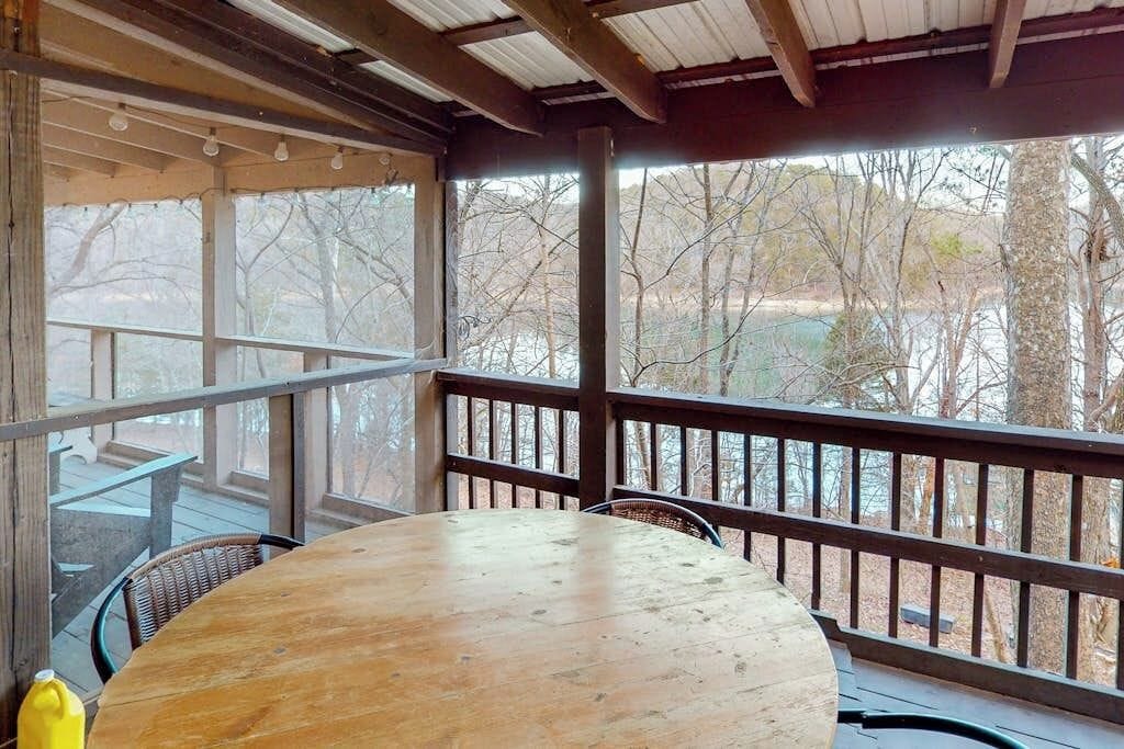 An outdoor wooden deck with a round table and chairs, a railing, and a view of trees and a body of water.
