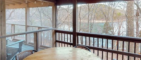 An outdoor wooden deck with a round table and chairs, a railing, and a view of trees and a body of water.