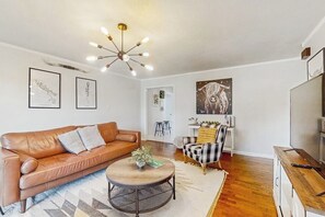 A living room with a brown leather sofa, a round coffee table, a plaid armchair, and a modern chandelier. - Large 3BR - Balcony - Firepit - Garage Parking (Bentonville)