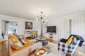 A living room with a sofa, a coffee table, a TV, and a chair. - Large 3BR - Balcony - Firepit - Garage Parking (Bentonville)