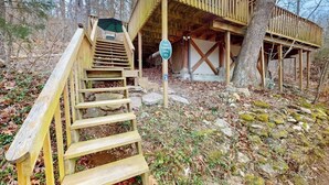 Eine Holzhütte mit einer Holztreppe, die zu einem überdachten Eingang führt, umgeben von Bäumen und natürlicher Vegetation.