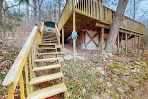 A cabin with a wooden staircase leading to a covered entrance, surrounded by trees and natural ground cover.