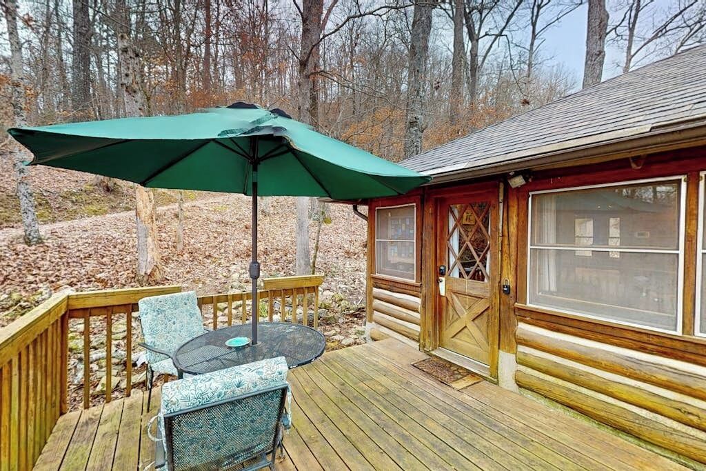 A wooden deck with a table and chairs, a green umbrella, and a cabin with a large window.