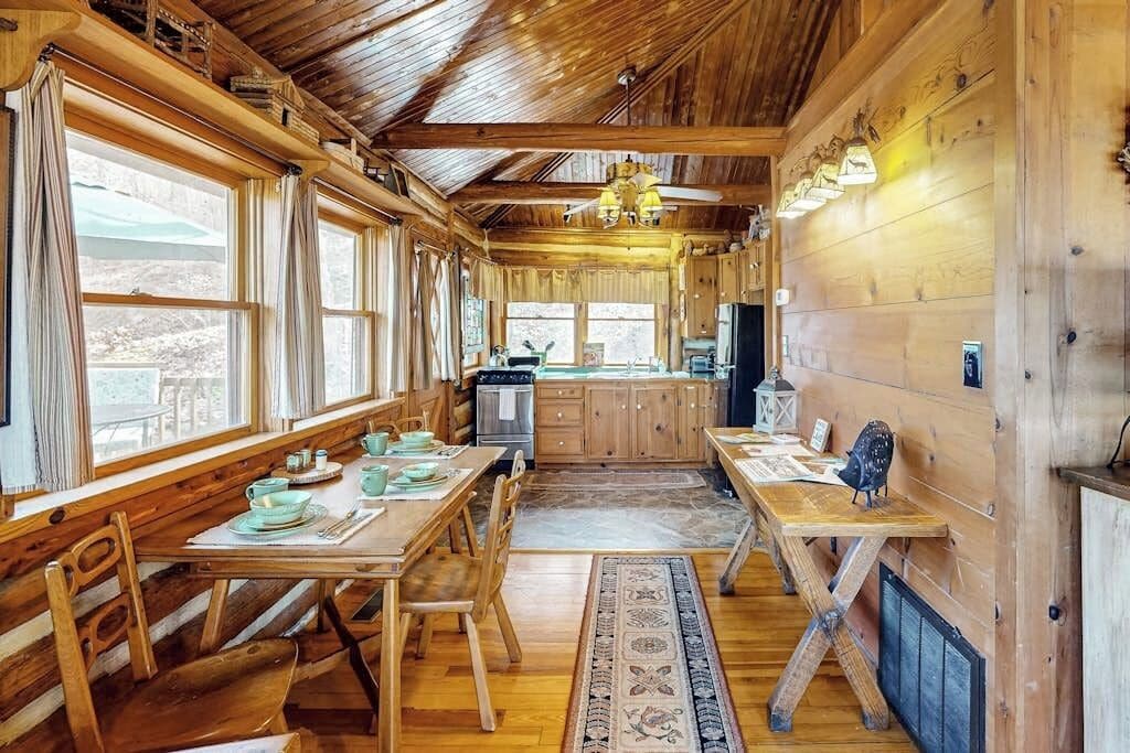 A wooden cabin interior with a dining area, kitchen, and large windows.
