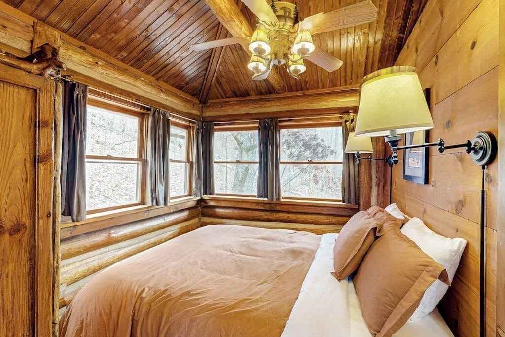 A log cabin bedroom with a bed, windows, a ceiling fan, and a wall-mounted lamp.