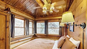 Ein Schlafzimmer in einer Blockhütte mit einem Bett, Fenstern, einem Deckenventilator und einer Wandlampe.