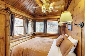 A log cabin bedroom with a bed, windows, a ceiling fan, and a wall-mounted lamp.