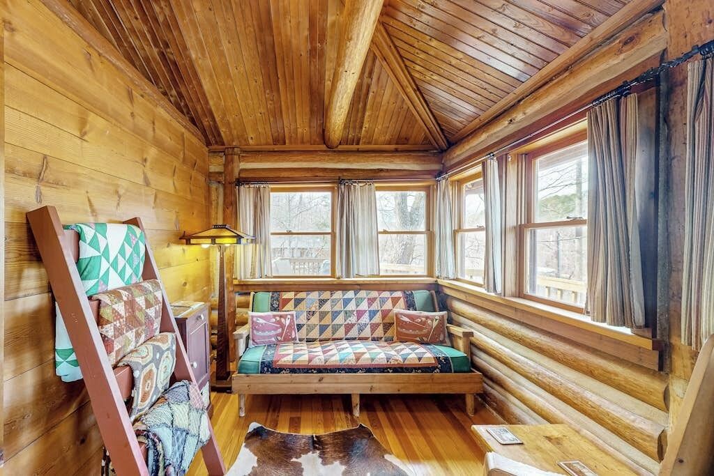 A log cabin interior with a wooden bench, large windows, and a lamp.