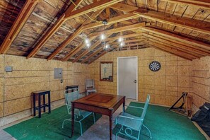 A rustic attic room with a wooden ceiling, a dartboard, a table, and metal chairs. - Whispering Trails | Bike-In/Bike-Out Retreat (Bentonville)