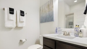 A bathroom with a toilet, a sink with toiletries, and towels hanging on a rack. - Stylish 2BR -  W&D - Downtown Location (Bentonville)