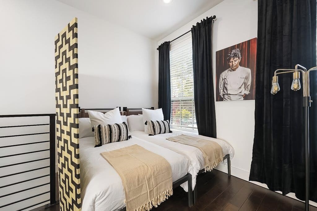 A bedroom with a bed, pillows, a bedside table, a window with curtains, and a wall-mounted light fixture.