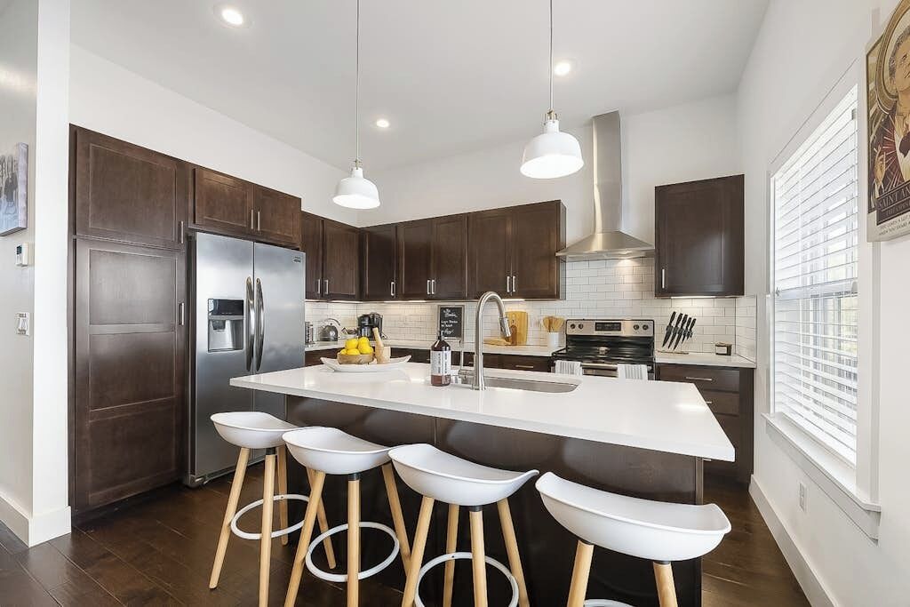 A modern kitchen with dark wooden cabinets, a stainless steel refrigerator, a white island with bar stools, and pendant lighting.