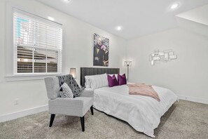 Ein Schlafzimmer mit einem Bett, einem Sessel, einem Fenster mit Jalousien, einem Wandbild und einer Stehlampe.
