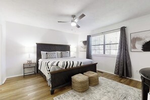 Una camera da letto con un letto, comodino, lampada, finestra con tende e una ventola a soffitto.