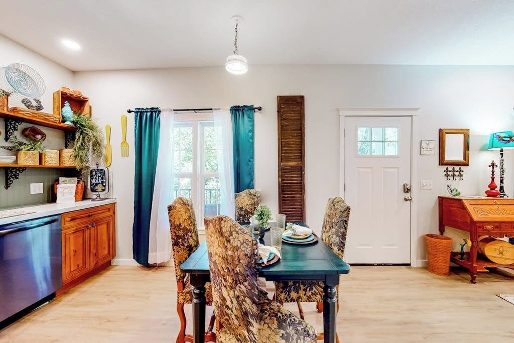 A dining area with a table set for four, surrounded by patterned chairs. A wooden cabinet with decorative items is to the left, and a door with a window is visible in the background.