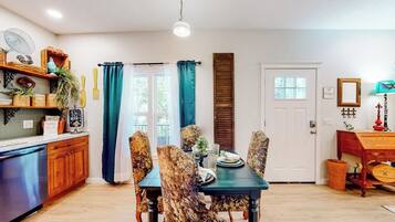 A dining area with a table set for four, surrounded by patterned chairs. A wooden cabinet with decorative items is to the left, and a door with a window is visible in the background.