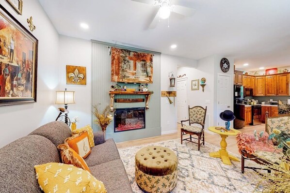 A living room with a sofa, ottoman, and chairs, a fireplace, and a kitchen in the background.