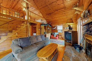 Ein Holzblockhaus-Interieur mit einer Couch, einem Fernseher, einem Kamin und einer Treppe.