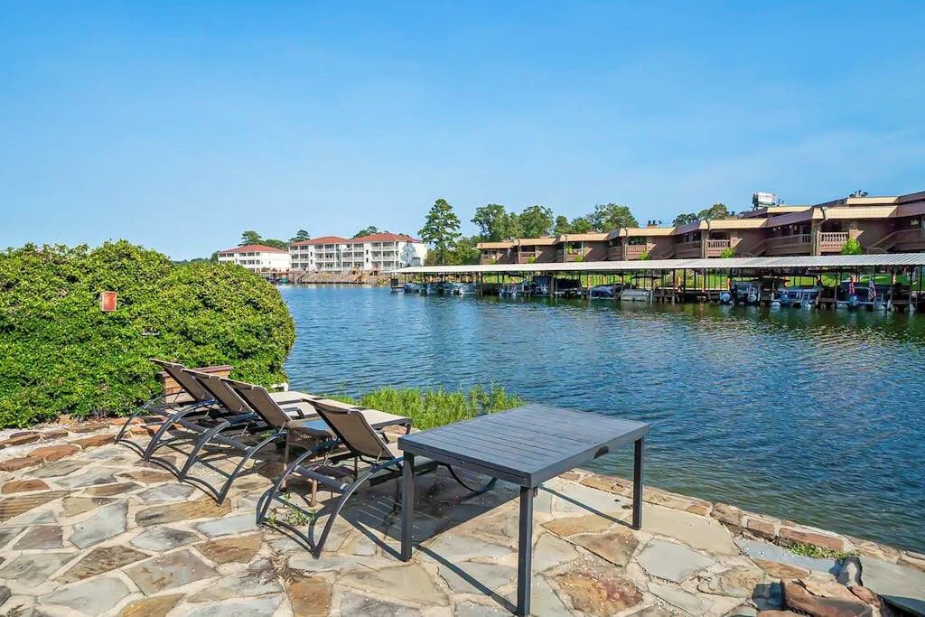 A waterfront patio with lounge chairs and a table, overlooking a calm body of water with buildings in the background.