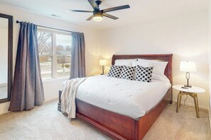 Une chambre à coucher avec un grand lit, deux lampes de chevet, un ventilateur de plafond et une vue sur l’extérieur par une fenêtre avec des rideaux.