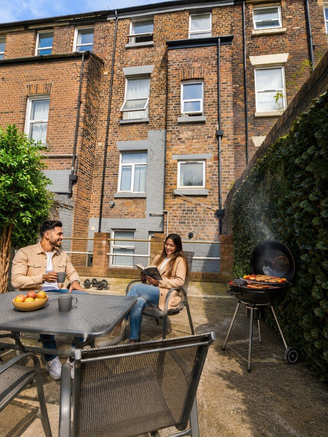 A brick building with multiple windows, a barbecue grill, and a table with fruits and a book.
