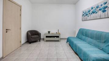 Une pièce avec une porte en bois, un fauteuil gris, un canapé turquoise, une petite table avec une plante et un tableau mural.
