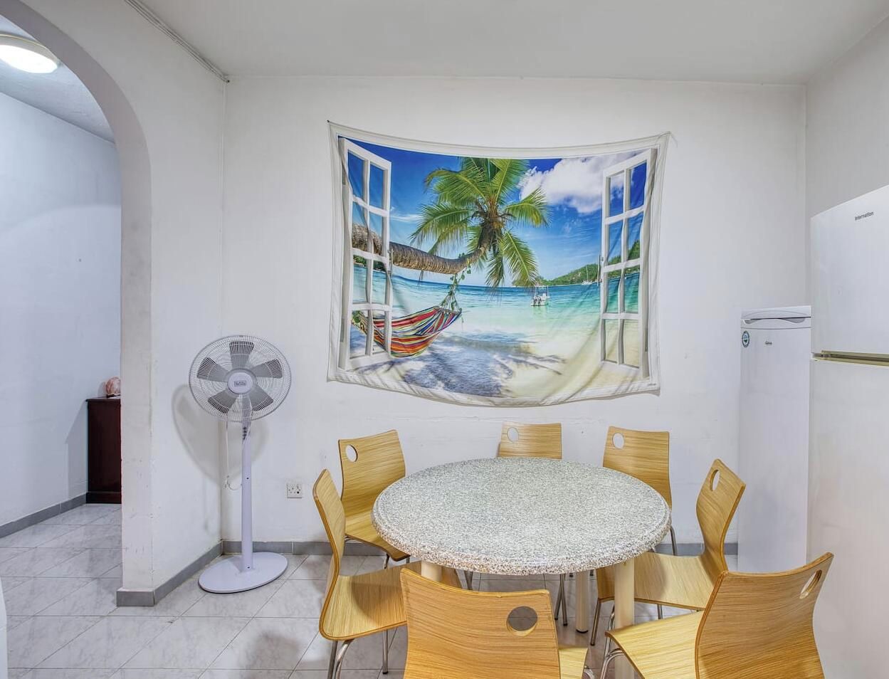 Un coin repas avec une table ronde et six chaises, un ventilateur sur pied et une fresque murale représentant une scène de plage.