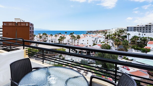 A balcony with a table and chairs, overlooking a coastal town with buildings and palm trees.