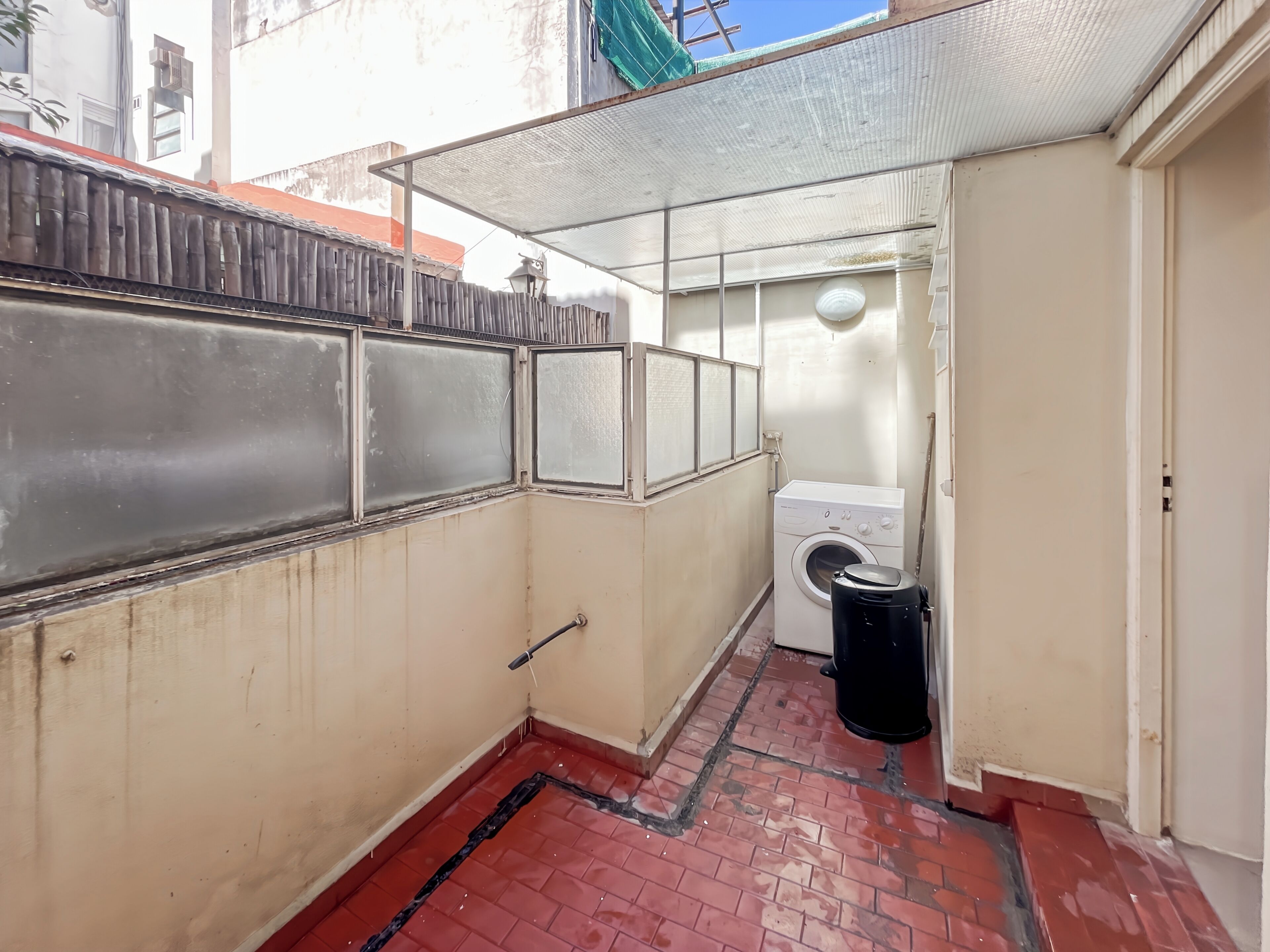 A compact laundry room with a washing machine, a black trash bin, and a metal fence.