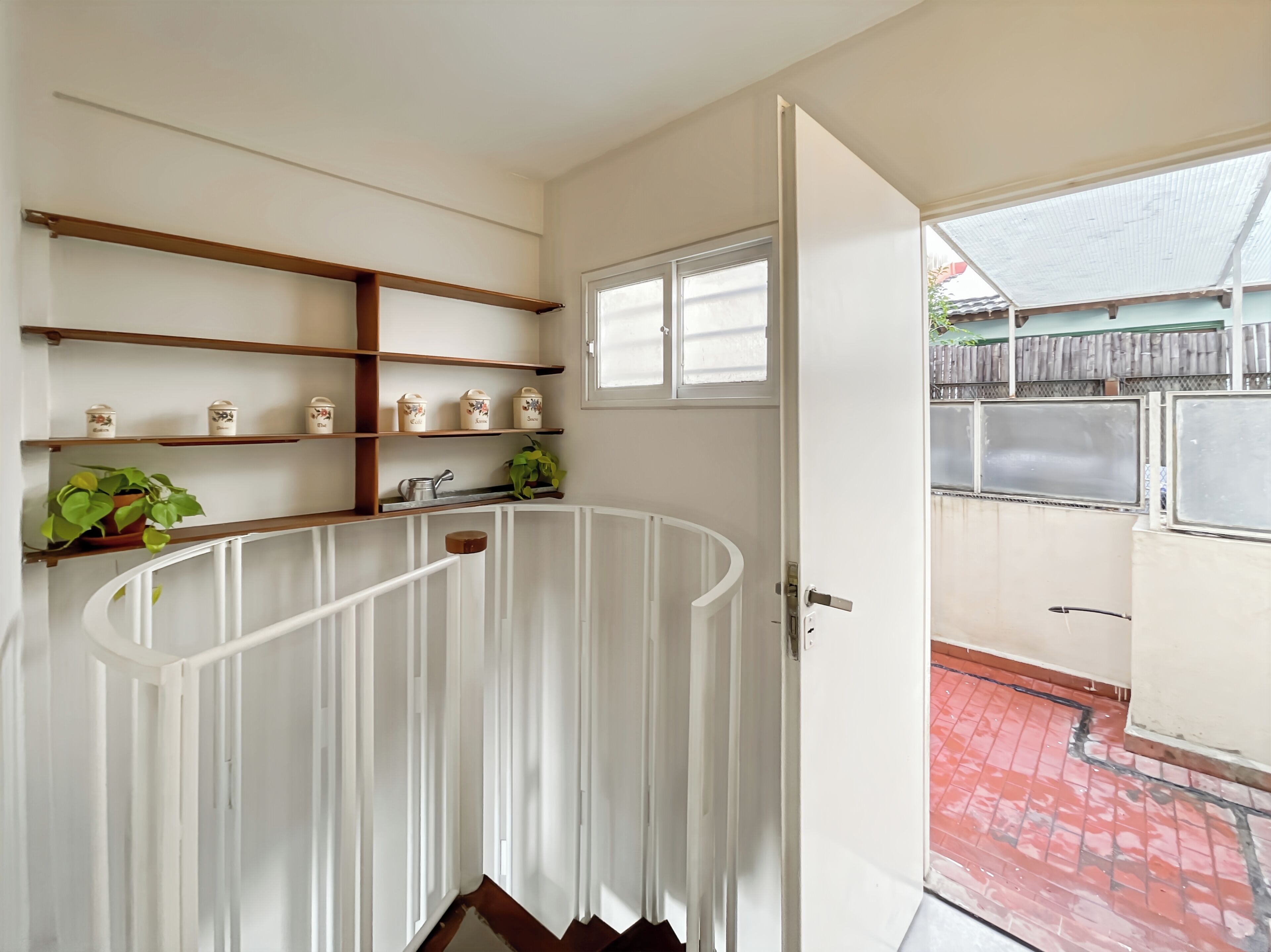 A room with a white railing, wooden shelves with jars, a window, and a door leading outside.