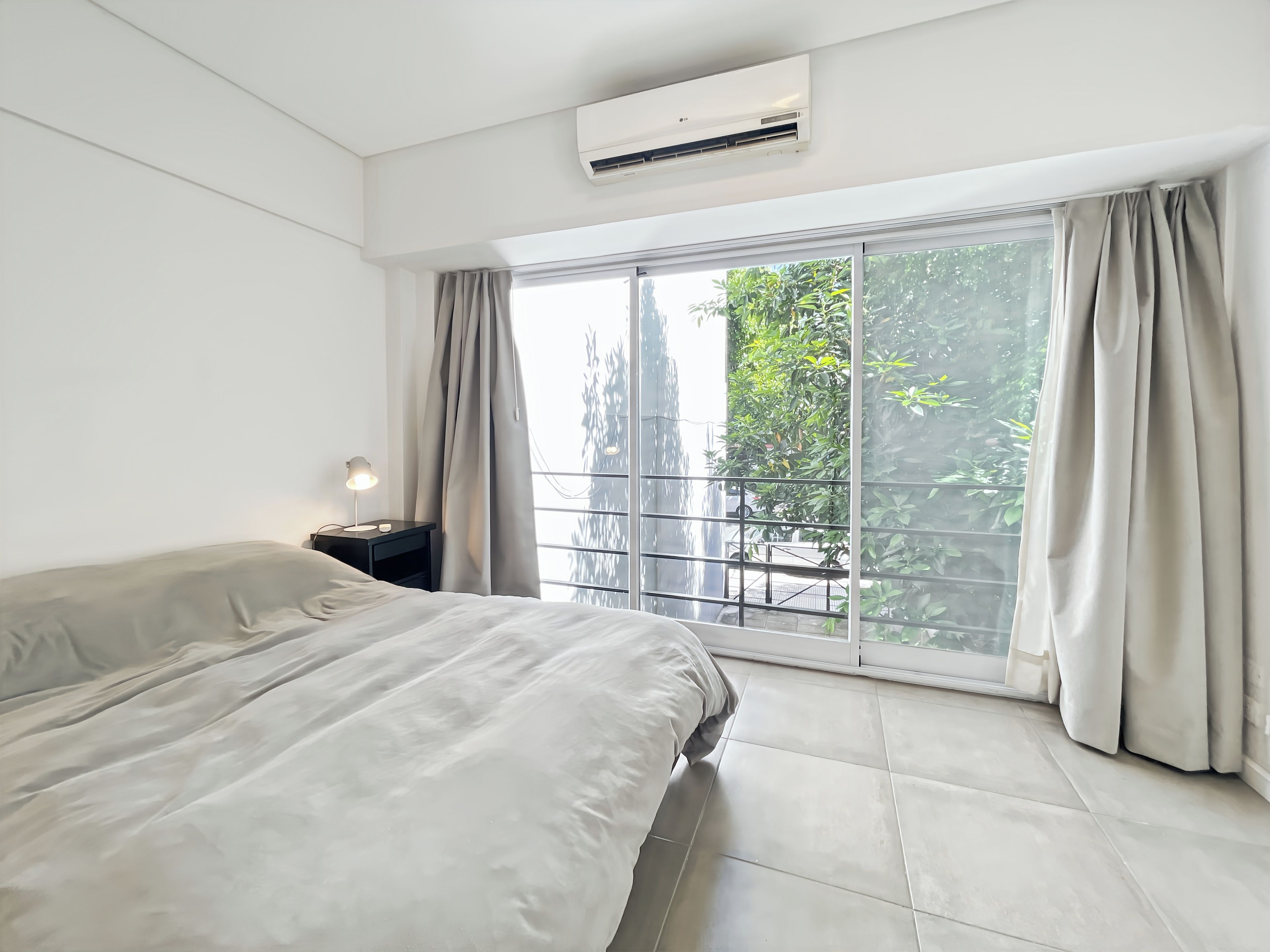 Un dormitorio con una cama, una mesita de noche, un equipo de aire acondicionado y un ventanal con cortinas.
