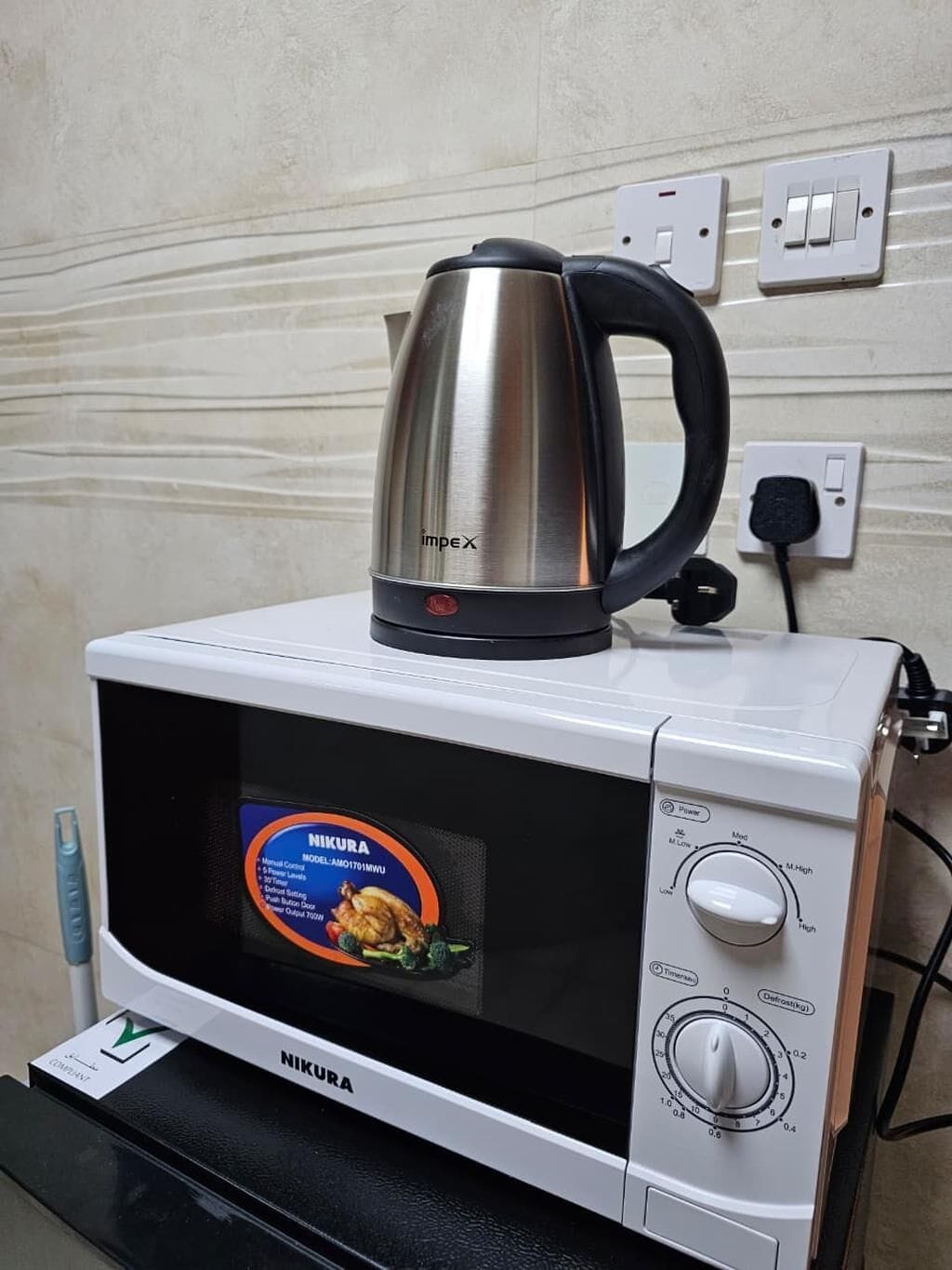 Een witte magnetron met een digitaal display, een waterkoker bovenop en een stopcontact aan de muur.