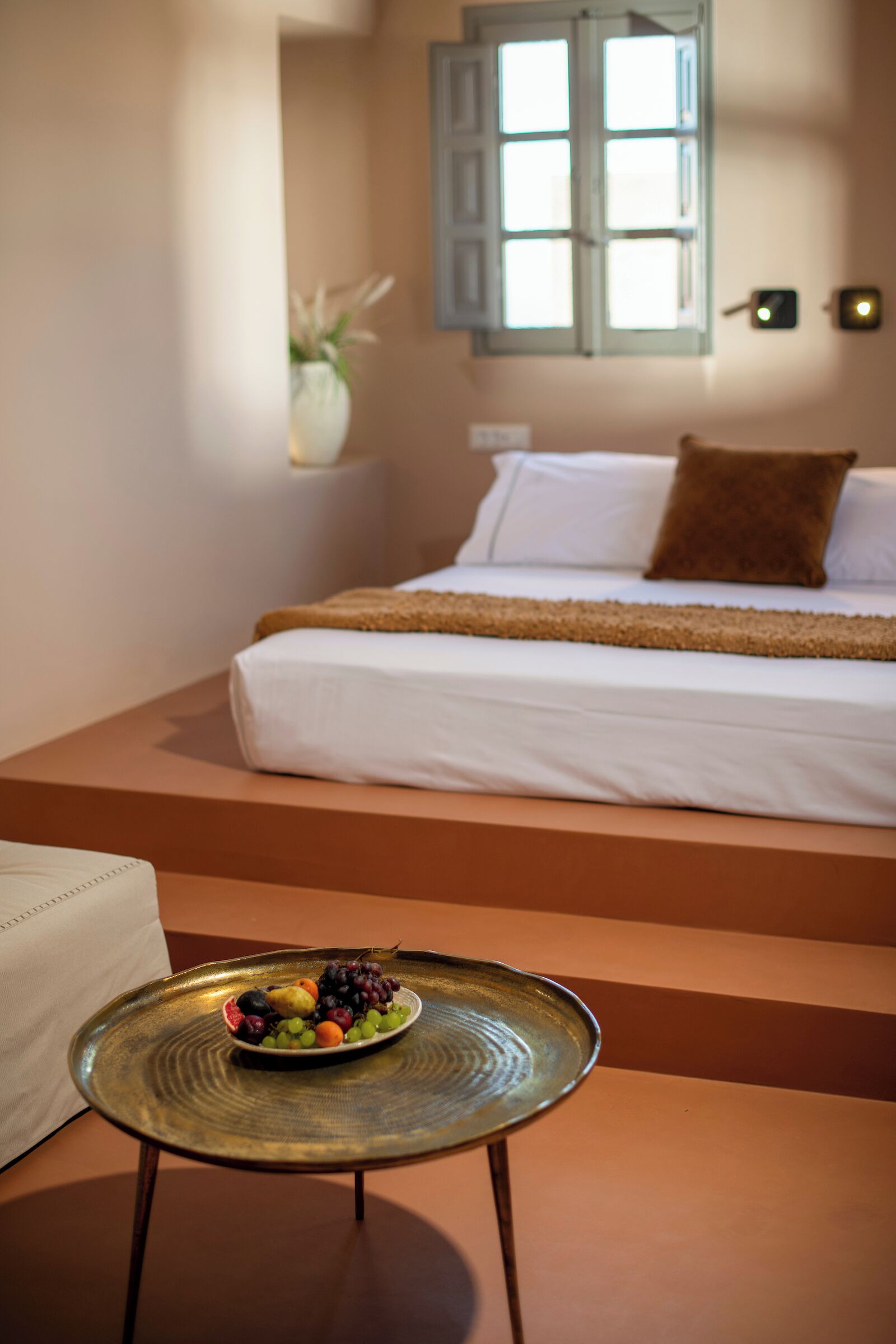 A bedroom with a bed, a bedside table with a fruit bowl, a window with shutters, and a wall-mounted light.