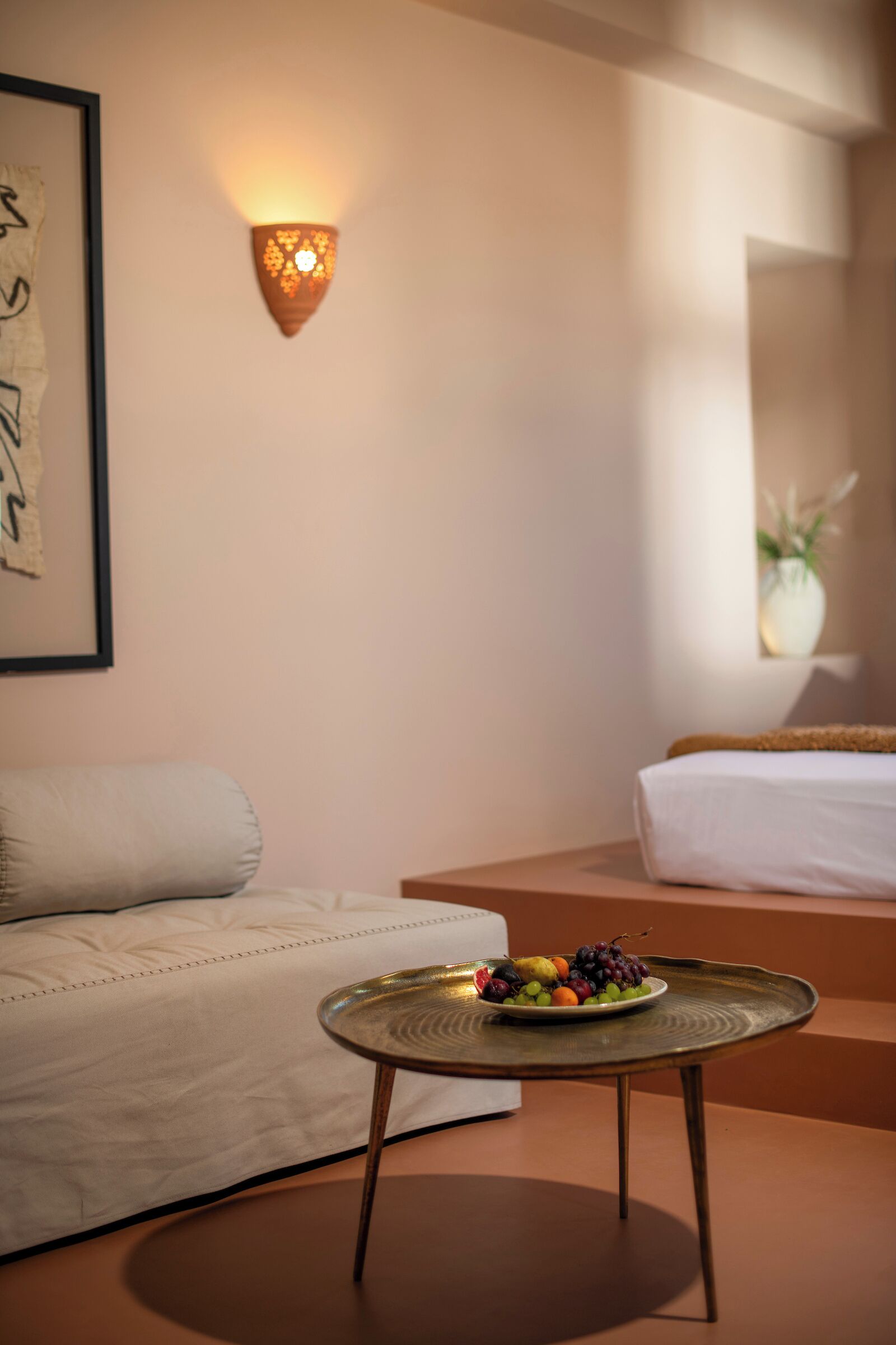 A hotel room with a bed, a small round table with a fruit plate, a wall-mounted light, and a framed artwork.