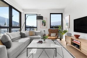 A modern living room with a sectional sofa, a coffee table, a side table, a television, and a large window with a mountain view. - Rest & Roam (Revelstoke)