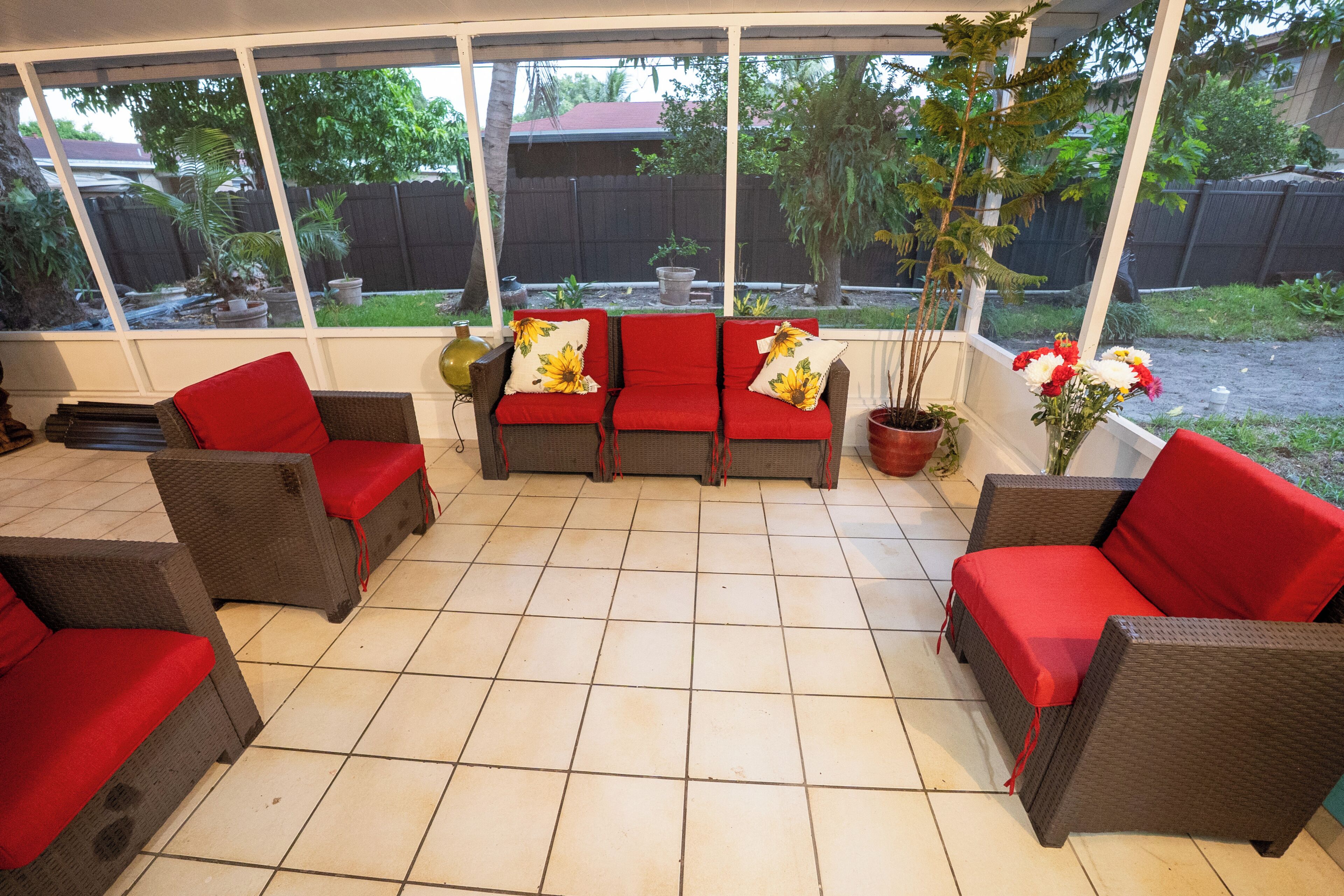 A covered patio area with wicker furniture and red cushions, a potted plant, and a view of a backyard.