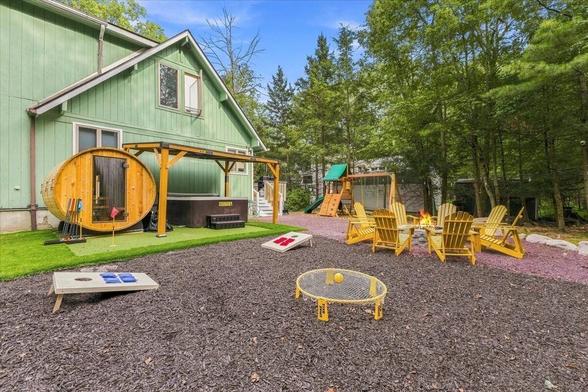 A green building with a covered outdoor area, a wooden barrel, Adirondack chairs, and a small fire pit.