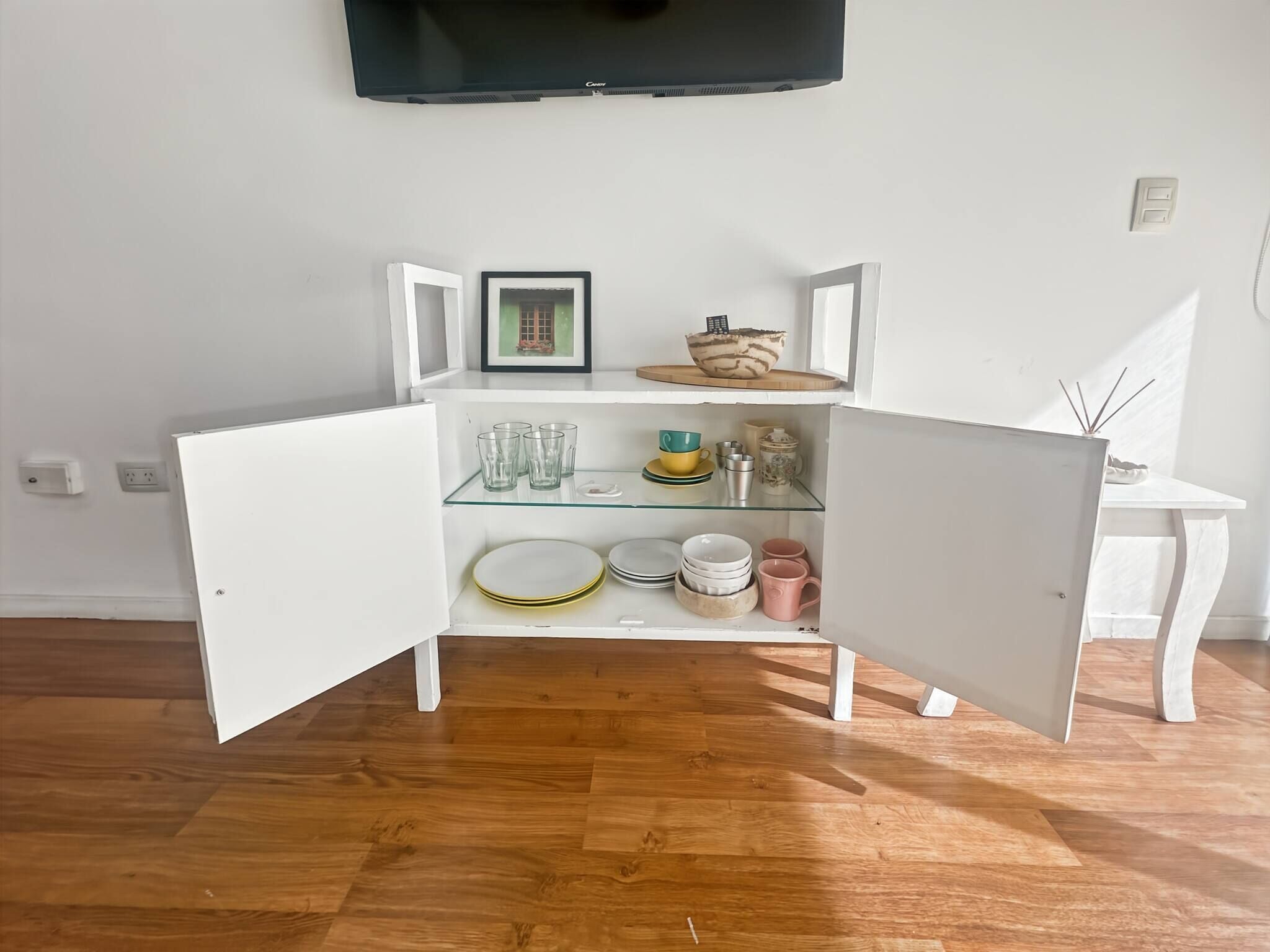 A room with a white cabinet with glass doors, a flat-screen TV mounted on the wall, and a wooden floor.