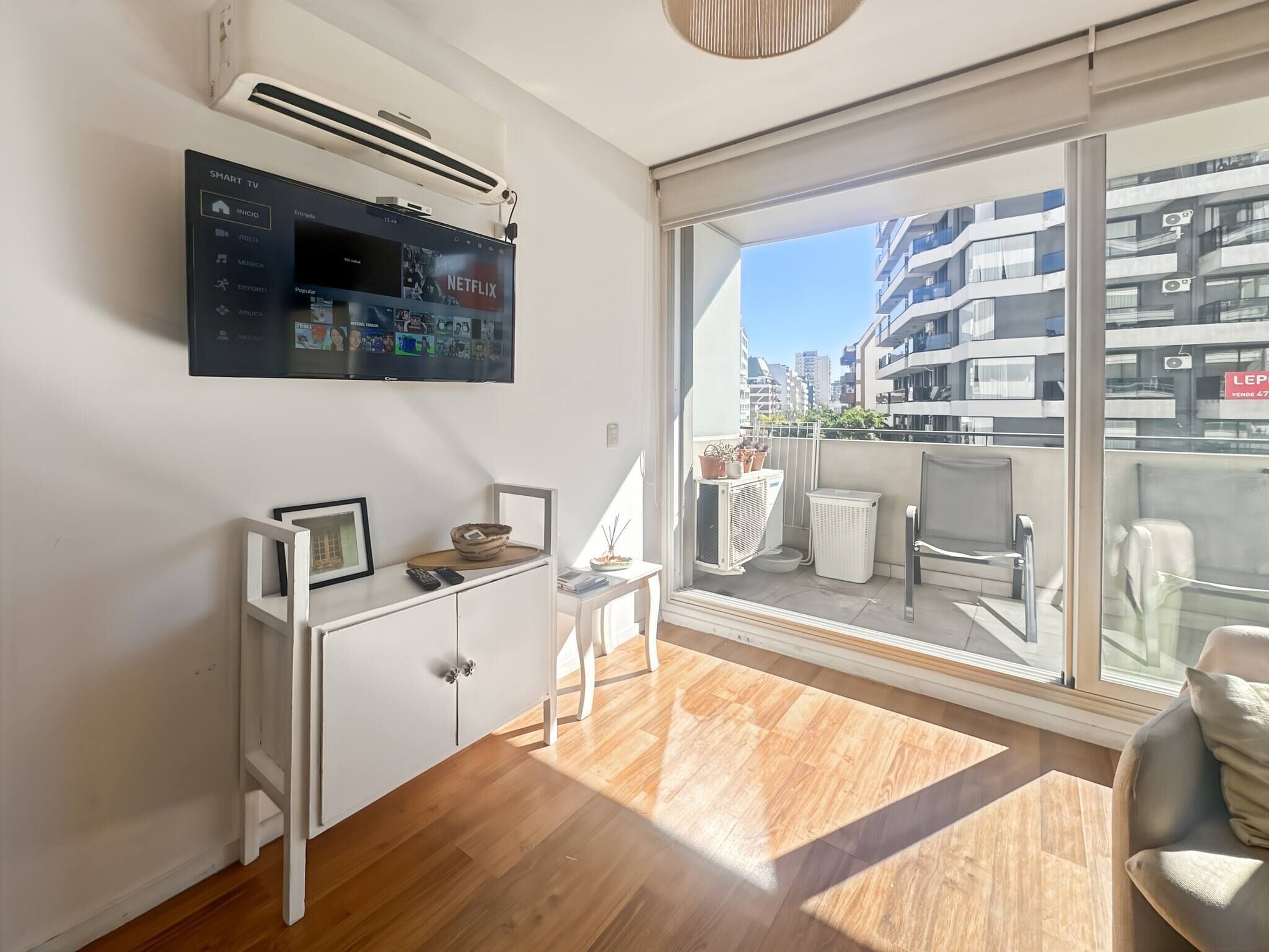 A modern apartment with a flat-screen TV, a white desk, a balcony with a view of the city, and a large window.