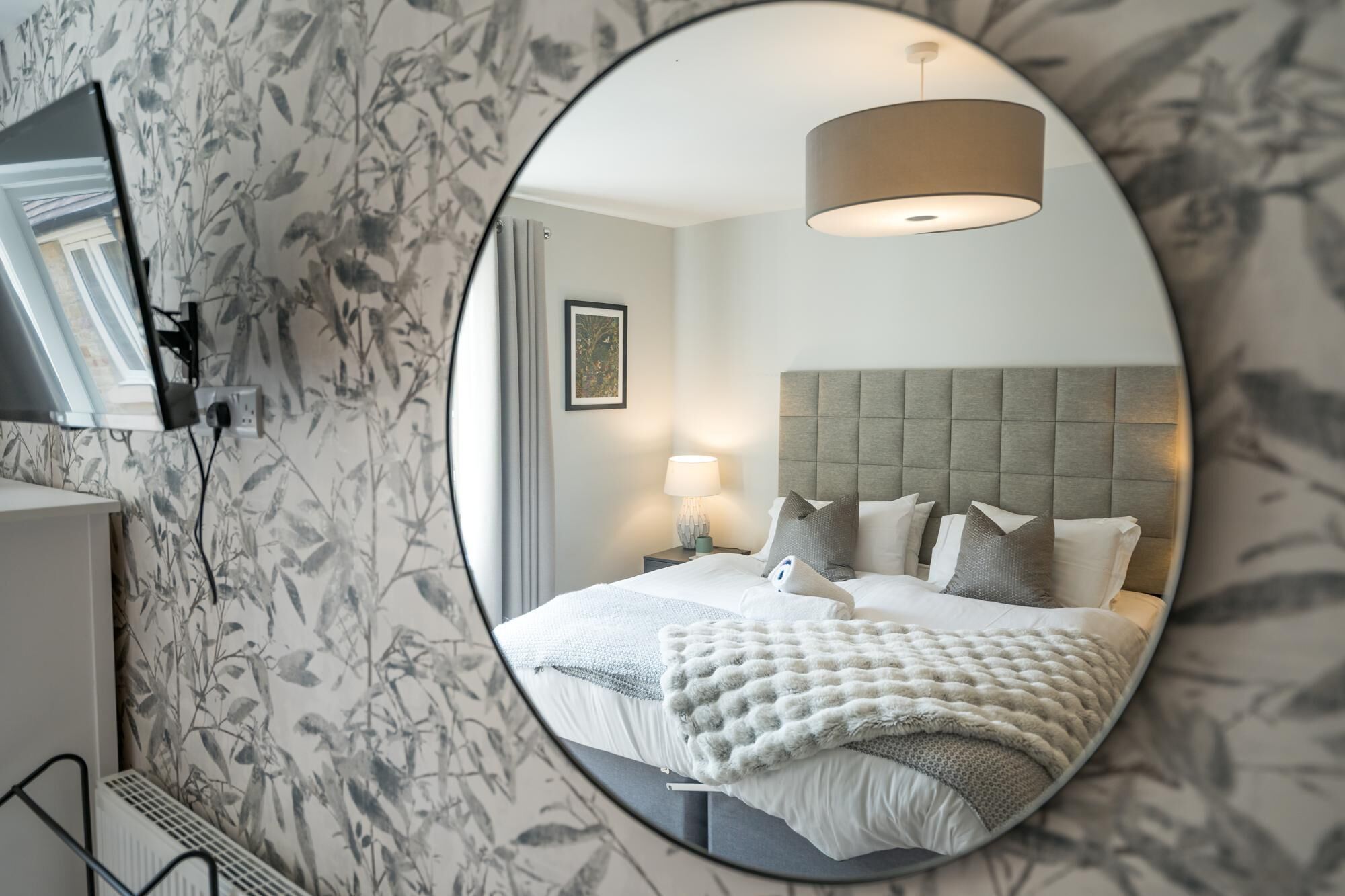 A circular mirror reflecting a bedroom with a bed, pillows, a bedside lamp, and a wall-mounted television.