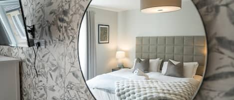 A circular mirror reflecting a bedroom with a bed, pillows, a bedside lamp, and a wall-mounted television.