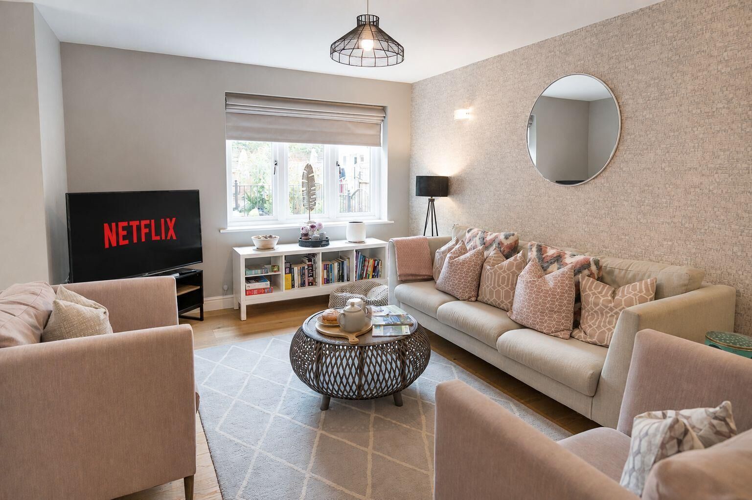 Un salon comprenant un canapé, des fauteuils, une table basse, une bibliothèque, un téléviseur affichant le logo « NETFLIX », et une fenêtre avec des stores.