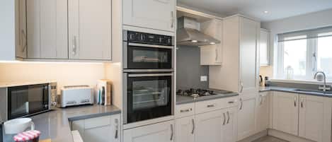Une cuisine moderne avec des meubles blancs, un four encastré, un four à micro-ondes, une cuisinière à gaz et une fenêtre laissant entrer la lumière naturelle.