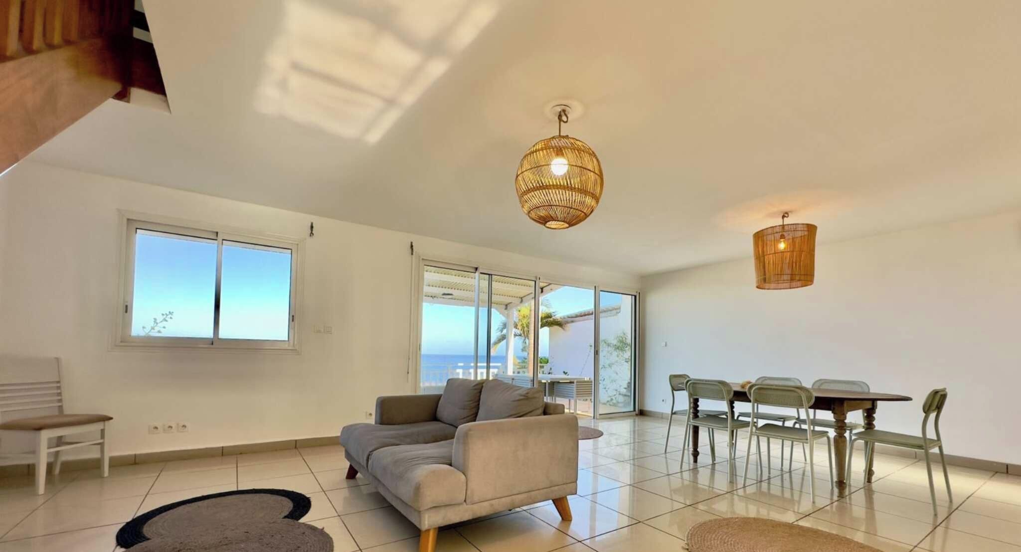 A spacious living area with a sofa, dining table, and chairs, large windows, and a view of the sea.